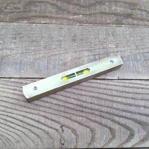 May include: A vintage brass level tool, rectangular with rounded ends. It features a clear glass vial with yellow liquid and a bubble. Two screws secure the top. The tool is on a weathered wooden surface. The level is approximately 20 cm long.