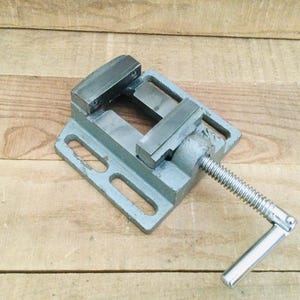 May include: A gray metal vise with a silver handle and screw mechanism. The vise has a square base with two rectangular openings and two metal jaws. The vise is designed to hold objects securely. The background is a wooden surface.