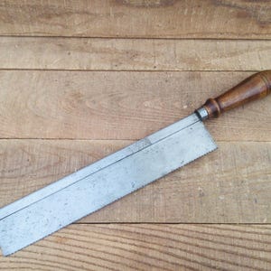 Peut inclure: Scie à main ancienne avec une poignée en bois et une lame en métal. La scie a une poignée marron foncé et une lame argentée avec des dents sur un bord. La scie est posée sur une surface en bois.