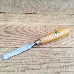 May include: A wood chisel with a light-colored wooden handle and a metal blade. The handle has a brass ferrule and a label with the number "115". The chisel is lying on a wooden surface.