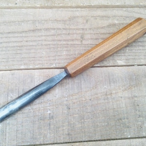 May include: A woodworking chisel with a wooden handle and a metal blade. The handle is a light brown colour and has an octagonal shape. The blade is a silver colour and has a curved end. The chisel is lying on a wooden surface.