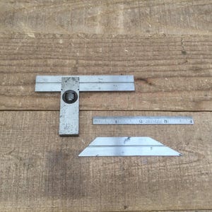 May include: A set of vintage metal tools, including a combination square, a small ruler, and a bevel gauge, displayed on a weathered wooden surface. The tools show signs of wear, indicating they are well-used.