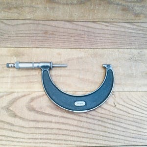 May include: A silver and black micrometer, a precision measuring instrument, rests on a wooden surface. The tool has a C-shaped frame with a black finish and a rotating barrel for fine adjustments. The micrometer is used to measure small distances.
