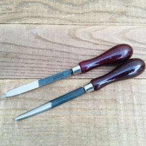 May include: Two metal files with dark burgundy wooden handles and silver ferrules are displayed on a wooden surface. One file is flat and rectangular, the other is round and tapered. The tools appear to be in good condition.