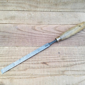 May include: A metal wood chisel featuring a long, flat, rectangular blade with a textured, weathered surface and a straight cutting edge. The tool includes a cylindrical wooden handle with a visible grain pattern and a metal ferrule at the base.