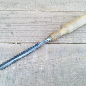 Peut inclure: Un ciseau à bois avec un manche en bois et une lame en métal. Le manche est en bois clair avec une forme cylindrique et une prise texturée. La lame est en métal foncé et se rétrécit en un bord plat. Le ciseau est posé sur une surface en bois.