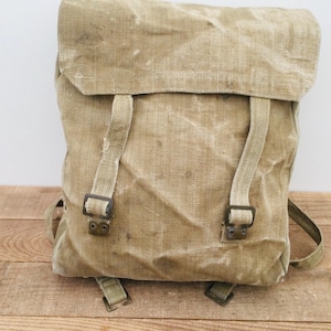 May include: A tan-coloured vintage backpack with a flap closure and adjustable straps secured by metal buckles. The bag is made from a robust, textured fabric, suggesting a durable design. The backpack is suitable for outdoor activities and measures approximately 40cm in height.