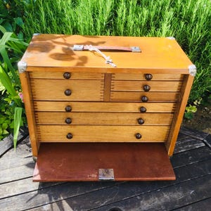 May include: A wooden chest with a top handle and metal corner reinforcements. The front features two small upper drawers and three full-width lower drawers, all with dark round knobs. A hinged wooden panel folds down at the base of the chest.