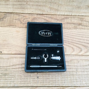May include: An open, black, felt-lined case containing precision measuring tools. The case features the logo "M&W" in white. The tools include a micrometer, a small wrench, and other components, all set against a wooden background.
