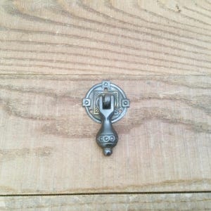 May include: A decorative, antique-style drawer pull with a dark gray finish. The pull features a circular backplate with a square design and a teardrop-shaped handle. The hardware is mounted on a light brown wooden surface.