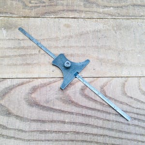 May include: A vintage depth gauge tool made of metal, resting on a wooden surface. The tool has a flat, star-shaped body with a sliding ruler and a screw for adjustment. The ruler is marked with measurements.