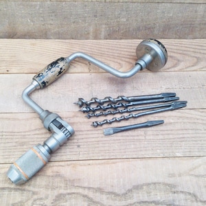 May include: A vintage hand drill with a silver finish and a wooden handle, accompanied by several drill bits. The drill bits are various sizes and shapes, suitable for different drilling tasks. The tools are arranged on a wooden surface.