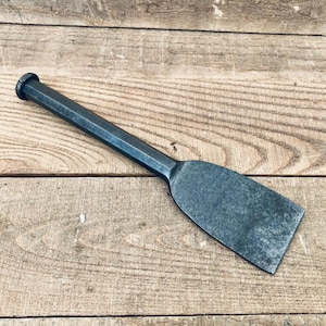 May include: A dark grey metal cold chisel featuring a long, hexagonal-shaped handle that tapers into a wide, flat, bevelled blade edge. The tool has a solid, industrial appearance with a uniform matte finish across its entire surface.