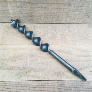 May include: A vintage auger drill bit, made of dark metal, lies on a wooden surface. The drill bit features a spiral design and a pointed tip. This tool is used for boring holes in wood.