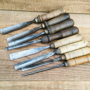 May include: A collection of seven vintage wood chisels arranged diagonally. Each tool features a weathered metal blade of varying widths and shapes, paired with a cylindrical wooden handle showing signs of age, wear, and natural wood grain patterns.
