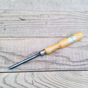 May include: A woodworking chisel with a light brown wooden handle and a black metal blade. The chisel has a metal ferrule where the blade meets the handle. The handle has a small, circular label. The tool is lying on a wooden surface.
