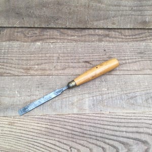 May include: A wood chisel featuring a long, flat, metallic blade with a beveled edge and a smooth, light-coloured wooden handle. The tool includes a small metal ferrule connecting the blade to the handle, showing signs of wear and age.