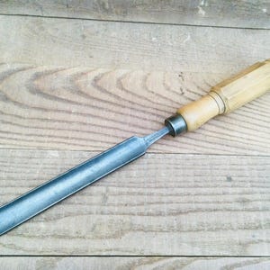 May include: A woodworking chisel with a wooden handle and a metal blade. The chisel is lying on a wooden surface. The handle is light brown and the blade is a silver color.