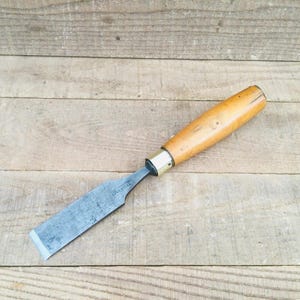 May include: A woodworking chisel with a wooden handle and a metal blade. The handle is light brown, and the blade is silver. The chisel is lying on a wooden surface.
