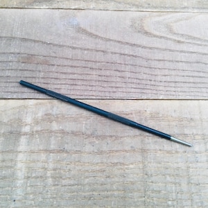 May include: A dark blue awl with a textured handle and a silver tip, lying on a weathered wooden surface. The awl is a slender, pointed tool, probably used for piercing or marking materials.