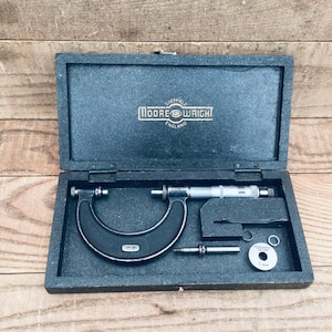 May include: An open, dark gray wooden box containing a black micrometer, a silver cylindrical tool, and several small metal pieces. The box lid has the text "MOORE & WRIGHT ENGLAND" in gold lettering. The tools are set against a dark gray interior.