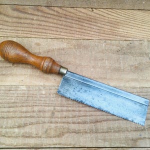 Peut inclure: Une scie à main vintage avec un manche en bois et une lame en métal. Le manche est de couleur brun chaud et la lame présente des signes d'usure. La scie est conçue pour la menuiserie fine et repose sur une surface en bois.
