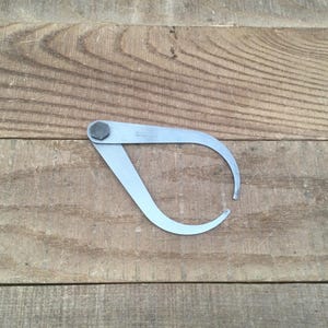 May include: A silver-colored outside caliper tool with a hexagonal bolt at the top. The tool has curved arms that extend downward. The tool is lying on a wooden surface with horizontal planks.