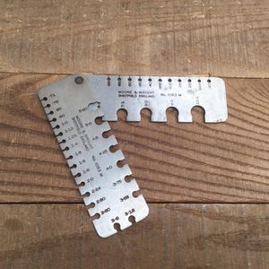 May include: Two vintage metal feeler gauges, one L-shaped and one straight, with measurement markings. The gauges are silver-colored and lie on a wooden surface. The text "MOORE & WRIGHT SHEFFIELD ENGLAND" is visible.
