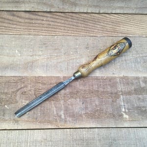 May include: A woodworking chisel with a wooden handle and a metal blade. The handle has a decorative design and a black end cap. The chisel is lying on a wooden surface.