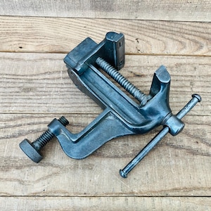 May include: A dark gray metal bench vise featuring a threaded screw mechanism, a sliding jaw, and a rotating handle. The tool includes a secondary threaded clamp at the base with a knurled knob for securing the device to a flat surface.