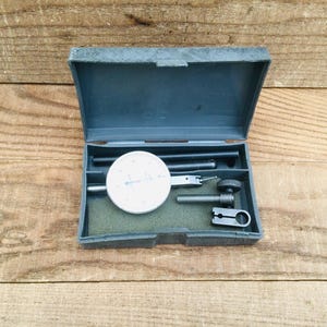 May include: An open, dark gray case containing a dial indicator and various measuring tools. The dial indicator has a white face with black markings and a needle. The tools are arranged in a green foam insert, set against a wooden background.