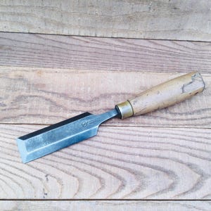 May include: A woodworking chisel with a wooden handle and a metal blade. The blade is angled and has a sharp edge. The handle is light brown and shows signs of wear. The chisel is lying on a wooden surface.