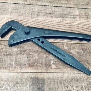 May include: A dark grey metal pipe wrench featuring a curved, serrated jaw and an adjustable pivot mechanism with multiple circular settings. The tool has a long, tapered handle and a secondary lever arm, with embossed text visible on the metal surface.