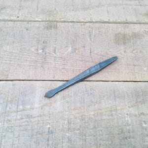 May include: A dark gray metal tool with a flat, angled tip and a textured surface. The tool is lying on a light brown wooden surface with visible grain.