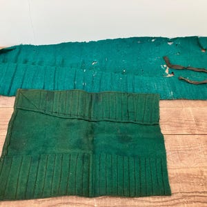 May include: Two green felt tool rolls featuring stitched vertical slots for organising small items. The larger roll is elongated with frayed edges and a black fabric tie, while the smaller roll is rectangular with multiple stitched compartments.
