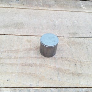 May include: A small, cylindrical metal container with a lid. The container is a dull silver colour and sits on a weathered wooden surface. The lid has a slightly lighter tone and appears to have embossed text or a design.