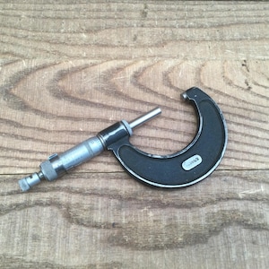May include: A black and silver micrometer tool, used for precision measurement. The tool is made of metal and has a curved frame with a measuring spindle. The micrometer is resting on a wooden surface.