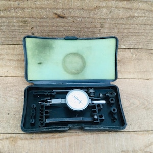 May include: A black dial indicator set in an open case. The dial has a white face with black markings and a needle. The case is black with a green foam insert. The set includes various attachments.