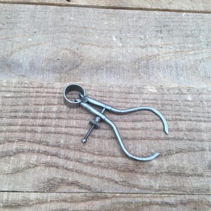 May include: A dark gray metal outside caliper tool featuring two curved legs connected by a circular pivot joint and an adjustable threaded screw mechanism with a small knob for precise measurement settings and locking the legs in place.