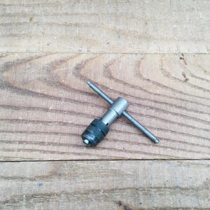 May include: A metal tap wrench tool with a knurled handle and a T-shaped design. The tool is silver and black, resting on a wooden surface. This tool is used for threading holes.