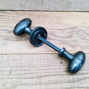 Peut inclure: Une poignée de porte noire de style ancien, avec une poignée ovale à chaque extrémité. La poignée est reliée à une plaque arrière circulaire par une tige carrée. La quincaillerie est en métal et a une finition vieillie.