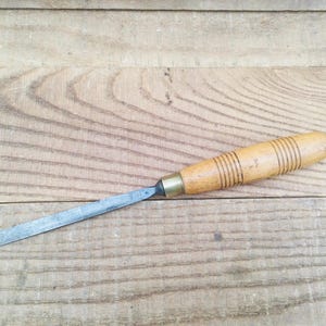 Peut inclure: Un ciseau à bois avec un manche en bois et une lame en métal. Le manche est brun clair avec des rainures circulaires brun foncé. La lame métallique est argentée et rectangulaire. L'outil est posé sur une surface en bois.