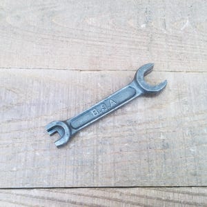 May include: A vintage, grey metal spanner with the letters "B.S.A." stamped on the handle. The spanner has two open ends of different sizes and rests on a light-coloured wooden surface.