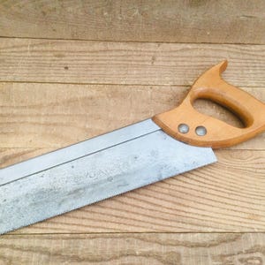 Peut inclure: Une scie à main avec une poignée en bois et une lame en métal. La poignée est brun clair et a une conception incurvée pour la prise en main. La lame est argentée et a un bord dentelé. La scie est posée sur une surface en bois.