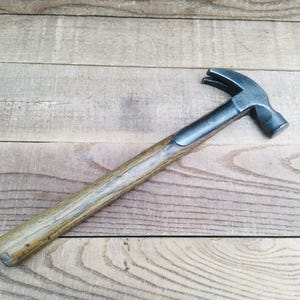 May include: A claw hammer featuring a dark grey metal head with a curved claw and a flat striking face. The tool has a long, straight wooden handle with a visible grain pattern, showing a smooth, worn texture along the length of the shaft.