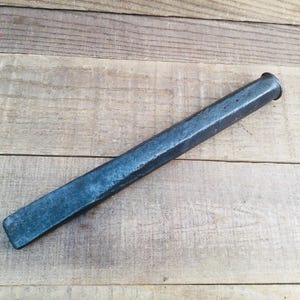 May include: A long, slender, dark gray metal tool with a square-shaped shaft and a slightly flared, rounded end. The surface of the metal appears textured and weathered, showing signs of use with a matte finish along its entire length.