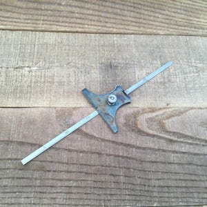 May include: A metal depth gauge tool featuring a flat, T-shaped base with a central locking screw and a thin, graduated ruler blade extending through the center. The tool has a weathered, dark grey metallic finish with visible numerical markings.