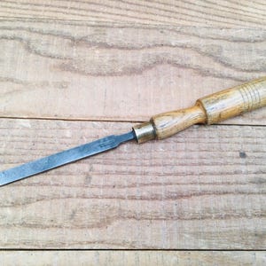 May include: A vintage wood chisel with a metal blade. The chisel has a light brown wooden handle with a brass ferrule. The blade is a rectangular shape and is made of steel. The tool is designed for woodworking and carpentry.