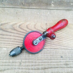 May include: A vintage hand drill with a red wooden handle and a red circular drill head. The drill bit is dark grey. The tool is set against a light brown wooden surface.