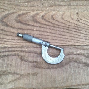 May include: A silver-colored micrometer tool, used for precise measurements, lies on a wooden surface. The tool has a curved frame with a measuring scale and a rotating thimble for adjustments. The micrometer is designed for measuring small distances.